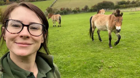 Marwell Zoo Hoofstock animal keeper Rhiannon Wolff taking a selfie in a green space at Marwell Zoo, with several Przewalski's horses walking in the background.