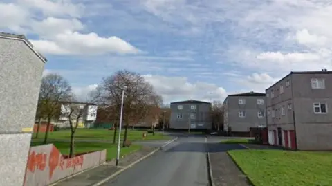 Street view of a residential estate with several grey three-storey apartment blocks on both sides of a quiet road. A grassy area and footpaths line the street, with a few leafless trees and a lamppost visible. The sky is partly cloudy with patches of blue, and no people or vehicles are present. Faint graffiti can be seen on a low wall in the left foreground.