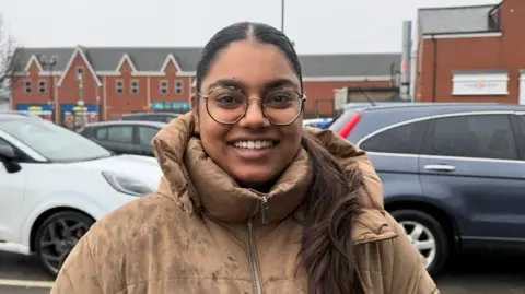 A woman smiling at the camera. She is wearing a brown jacket, glasses and her dark brown hair is tied back into a ponytail. She's standing in a car park and there is a modern-looking terrace of shops behind her. 