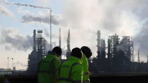 Three people in yellow high visibility jackets and black woollen hats walking in front of the smoking chimneys of the Lindsey Oil Refinery