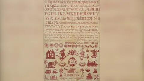 The sampler. It has sewn images, including an anchor, an elephant with some trees, the name Dora, a cenotaph cross, a cat, a sailing ship, the Prince of Wales' feathers.