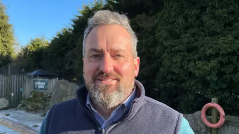 Simon Cox, a middle aged man with grey hair and a beard, smiles at the camera as he stands in the penguin enclosure at Birdland Park and Gardens in Gloucestershire on a sunny day. He is wearing a blue striped shirt and navy fleece gilet.