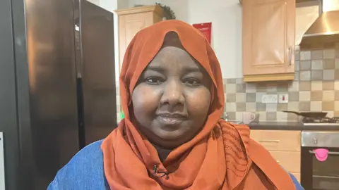 BBC Suha, wearing an orange head scarf and a denim shirt, with a kitchen in the background