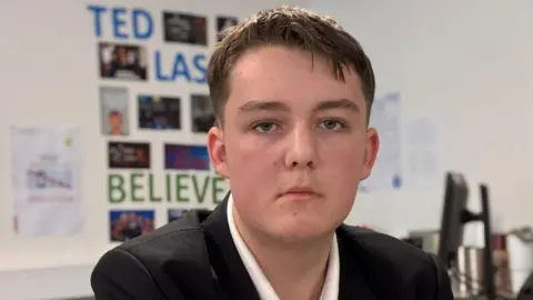 A boy with short brown hair. He is wearing a white shirt and black blazer and is looking at the camera with a serious expression.