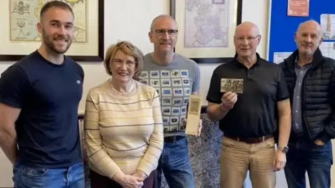 Philipp Cross A young man in his 20s wears a blue t-shirt and jeans, standing next to one elderly woman and three older men at a library. Two men in the middle are holding WW1 postcards