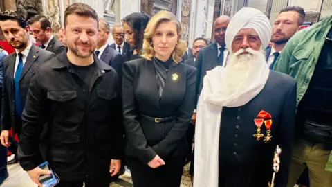 Bhai Sahib The Ukrainian president, Volodymyr Zelensky, a man with short dark hair, is wearing a black T-shirt under a black shirt. He is standing next to his wife, Olena Zelenska, who has shoulder-length blonde hair and is wearing a black suit. They are with a bearded man, Bhai Sahib, Professor Mohinder Singh, who is wearing a long black jacket two medals.