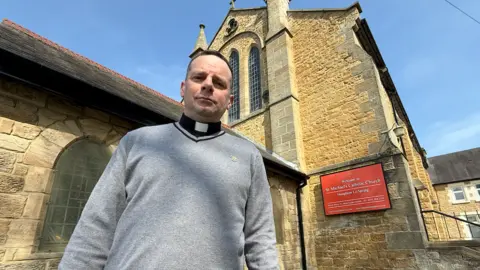 Father Marc Lyden-Smith wearing a grey jumper and a black and white vicar's collar stands in front of a church he looks after, which is yellow-bricked and has a red sign on it reading Welcome to St Michael's Catholic Church in Houghton-le-Spring