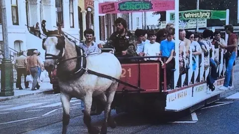 Isle of Man Transport A old photograph of an open top horse tram being driven by Peter Cannon and pulled by a white horse.