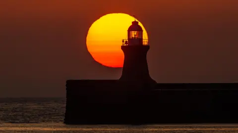 Weather Watchers/A Figment of your Imagination South Shields Lighthouse, Tyneside, silhouetted against a large, red sun. The sky is deep red and in the foreground is the sea.