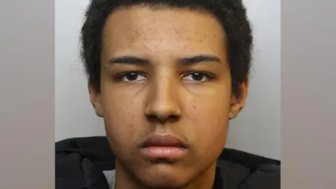 A mugshot of Romeo Dumbi. He has short black hair and some stubble on his upper lip. He is pictured looking directly at the camera with a blank expression.