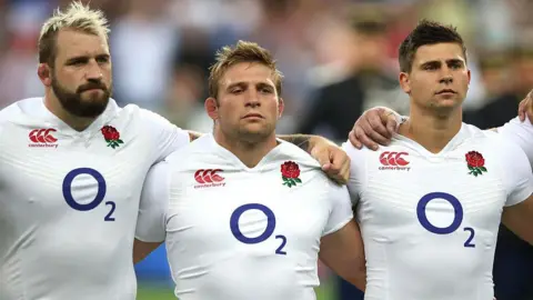Getty Images Three men in white England rugby kits stand with arms round shoulders and stare intently off camera. Left to right is a man with dyed blond spiky hair and a brown beard, a shorter man with blond hair and a slightly taller man with short brown hair.