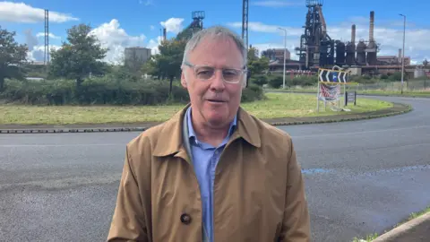 Alan Coombs wearing a brown jacket and blue shirt. He is standing in front of the steelworks in Port Talbot and is looking at the camera. 