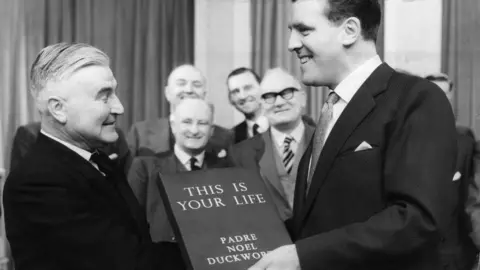 A black and white image showing Noel Duckworth being on This is Your Life in 1959. He is on the left, in profile, wearing a black suit with slicked back white hair. Eamonn Andrews is on the right, in a dark suit and smiling broadly while handing him the Book of his Life, which has This is your life Padre Noel Duckworth written on it. Behind them is a smiling group of suited men. 