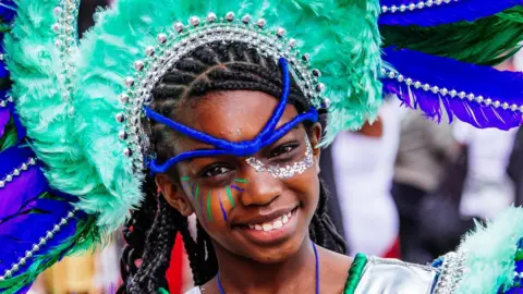 Aleksandra Warchol A child, dressed up for carnival, with make-up on her face, and a headdress on in blues and greens. She is smiling and looking straight at the camera.