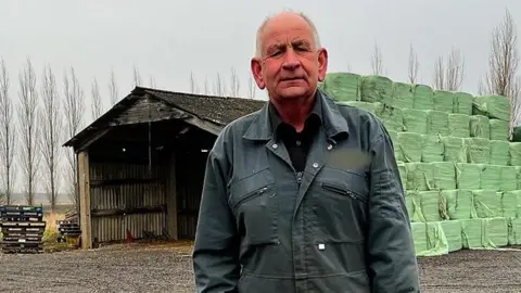 Councillor Julian Kirk wears dark blue overalls. He stands in front of a large barn and bales of animal forage wrapped in green plastic. 