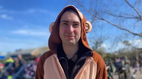 A man in a monkey onesie smiles for the camera. There are bikers behind him, out of focus.