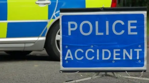 A close up of a blue rectangular sign, resting on tarmac, saying police accident in white letters. Behind it is a close-up of the rear view of a police car, marked with blue and yellow rectangles. 