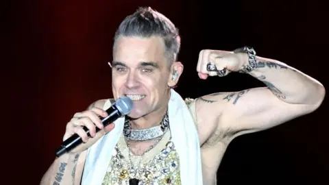 Robbie Williams sings into a microphone on stage. He smiles wearing a sparkly vest