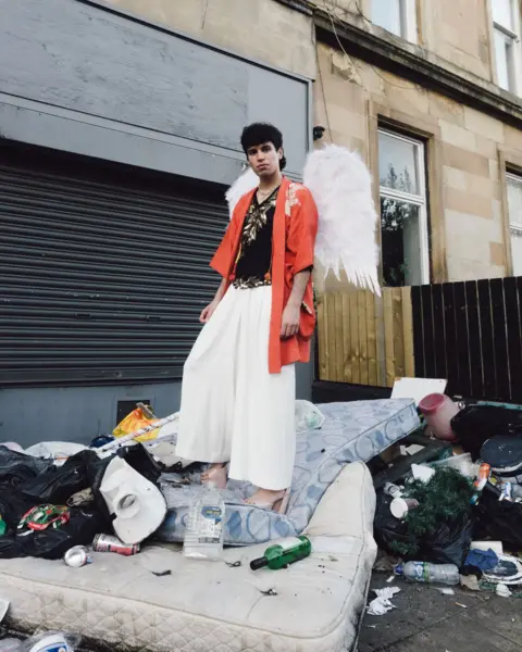 Island Records Jacob Alon, wearing a pair of white feathered wings, stands in a back yard that's been used for fly-tipping mattresses, old clothes and other household waste