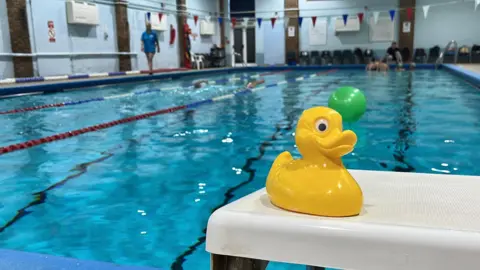 the centre's four lane 20 metre pool which has people swimming in it. On one of the diving podiums is a yellow rubber duck.