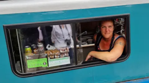 The Roving Canal Traders Association A woman with a tattoo on her arm leans out of a window of a teal boat. Through the window, cans of drinks, packaged snacks, and a food hygiene rating of five are visible. 