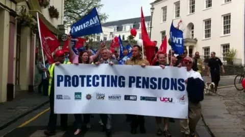 BBC A white banner with the words protecting pensions being held by a number of people at the front of a column of protestors holding banners. 