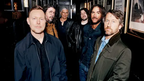 Six members of the Foo Fighters band stand side by side looking directly at the camera, hands in pockets.