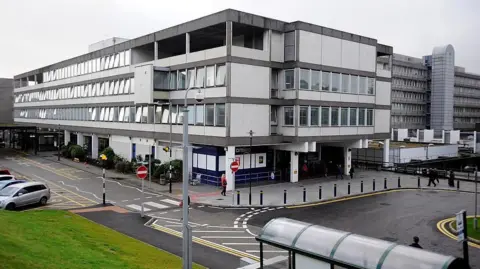 Grey buildings in the hospital complex. The buildings have multiple floors. There is a car park and a bus shelter.