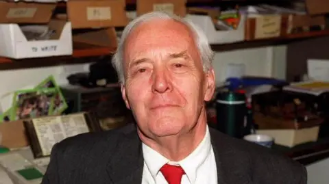 PA Media MP Tony Benn sits inside an office with boxes of paperwork behind him. He has short, grey hair and is wearing a black suit jacket with a white shirt and red tie.