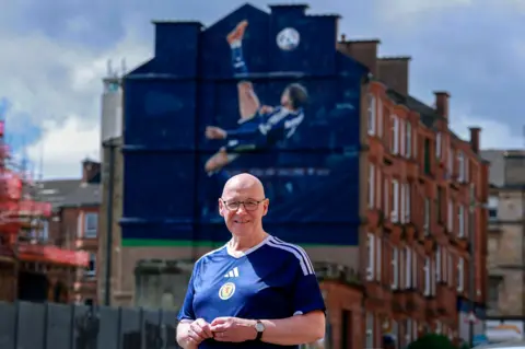 Getty Images A smiling First Minister John Swinney stands in front of a mural, on a building in Glasgow, featuring Scotland midfielder Scott McTominay scoring in the 4-2 victory over Denmark
