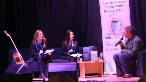 Gemma Kelly Two female school pupils sit on a couch carrying out an interview with Stuart Murdoch - a man with short hair, holding a microphone. 