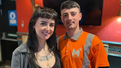 A woman with long dark hair, and a man with short dark hair, stand arm in arm smiling in a pub. The woman is wearing a grey jacket and black top. The man is wearing bright orange overalls.