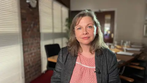 Tracy Daszkiewicz looks straight down the camera lens as she sits in an office. She has shoulder-length blonde hair and is wearing a pink patterned top under a grey blazer. 