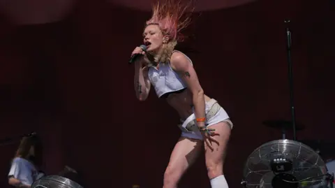 PA Media A woman wearing white shorts and a white crop top is singing into a microphone on stage with her hair sticking up, blown by fans.