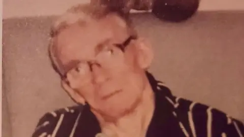An elderly man in a striped dressing gown, with grey hair and glasses looks at the camera. It is a faded, slightly blurred picture.