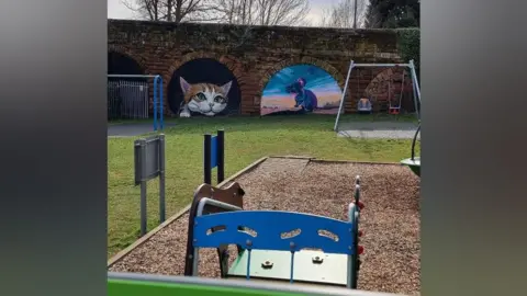 Tim Robottom The two murals in the brick arches are in the background. In the foreground is a play area with bark on the floor and a pair of swings. Other equipment is blue and green.