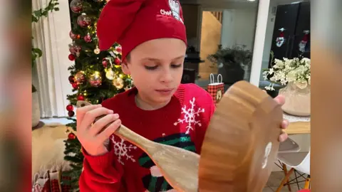 Joshie Harris pictured baking pies in his kitchen. He is holding a wooden spoon and bowl while wearing a red hat saying 'chef' and a red jumper with a white snowman on the front. In the background is a Christmas tree decorated with baubles. 