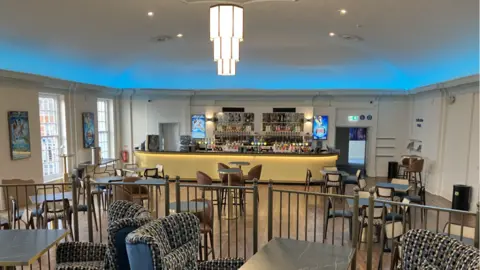 Shaun Whitmore/BBC A view of a bar taken from a seating area, showing tables and chairs and Art Deco style lighting 