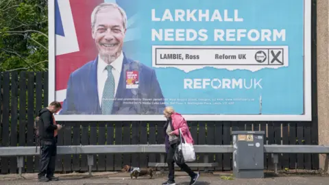 A large billboard image for reform featuring Nigel farage and a Union flag, two people walk in the foreground.