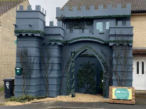 Grey fabricated castle structure on the driveway of a house, with towers on each corner and an arch-shaped gate at the front. Topiary runs around the frames of the doors and the division between the ground floor and second floor of the castle.