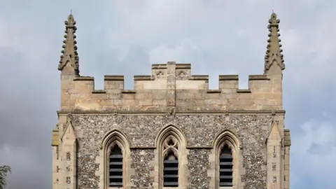 Historic England A close up of the top of a stone bell tower. The sky is cloudy behind it. 