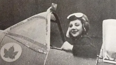 Candy Adkins A woman in flight gear. She is sat in the cockpit of a vintage plane. The image is black and white. 