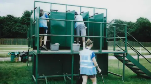 WaterAid A film photo showing three WaterAid volunteers wearing hi-vis blue branded vests. They are cleaning the toilet stalls on a green metal raised platform.