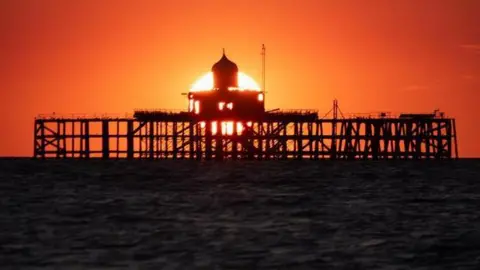 Omri Moxey A construction in the middle of the ocean with the sun going down behind it with an orange sunset 