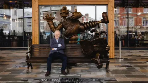 Preston City Council Nick Park sits on the bench that forms part of a statue of Wallace and Gromit. He wears a blue jacket, and smiles at the camera with his arms folded. The bronze-coloured statue includes Wallace, who stands behind the bench in "the wrong trousers". Gromit sits on the right side of the bench reading a newspaper.  