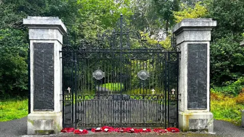 A set of memorial gates with a marble pillar either side. At the base of each post is a gap where there was once a plaque