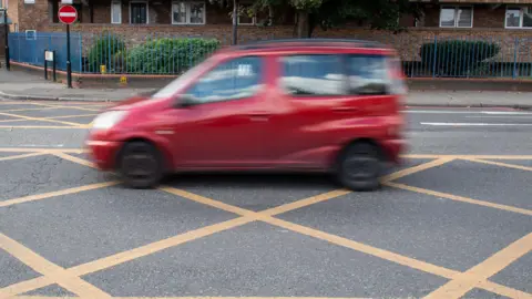 Small red car blurred out passing through a yellow box junction on a main road.