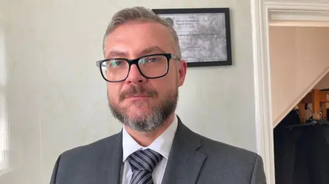 Michael McCulloch has short hair and a beard. He is wearing dark-framed glasses and a grey suit with a white shirt and a navy and grey striped tie.