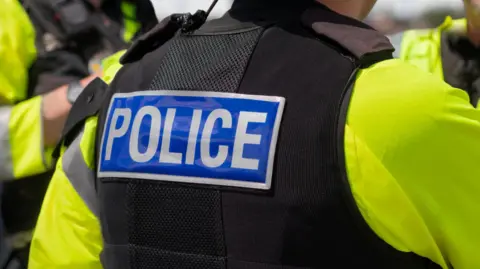The back of a police officer's jacket with "POLICE" written on it, as three police speak together. Their faces are all hidden.