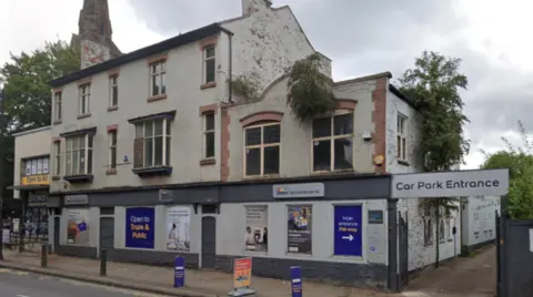 A three-storey building has the Dulux logo on the outside at the top of the ground floor. A road is on the bottom left of the image and a sign with the words car park entrance is on the right.